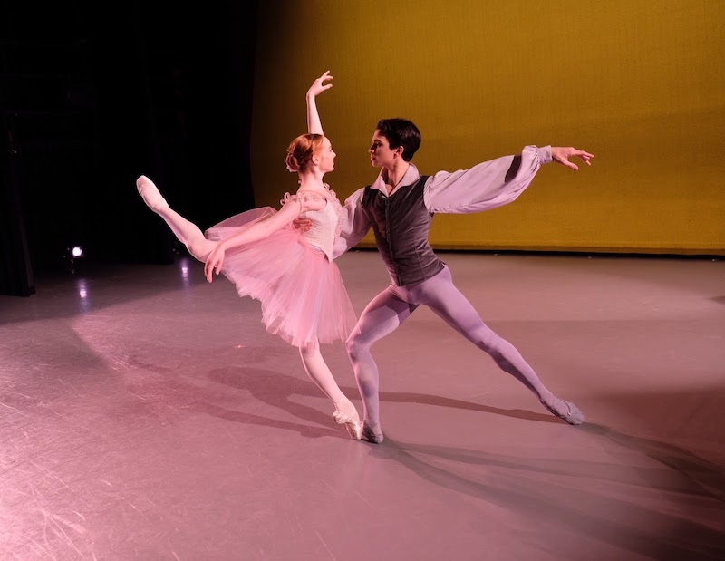 A woman in a pink tutu assumes a high attitude position en pointe that is slightly off kilter. Her male partner supports her back.